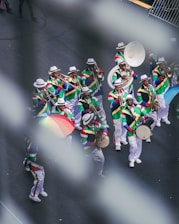 A vibrant marching band dressed in colorful, patterned costumes with white hats. They are playing various brass instruments like trombones, saxophones, and trumpets. One member holds a multicolored umbrella, adding to the lively scene on a city street.