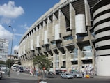A large, modern sports stadium with a curved roof and prominent architectural features dominates the scene. Surrounding the structure, people and several vehicles are visible, indicating a busy urban environment. Flags and banners suggest an event or gathering. Nearby, urban buildings and trees add to the bustling atmosphere.