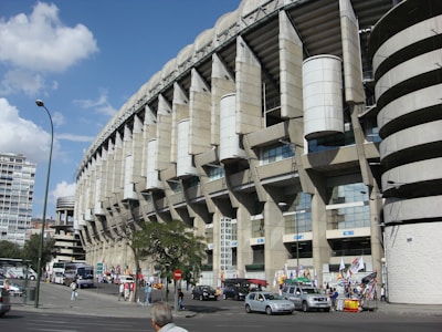 A large, modern sports stadium with a curved roof and prominent architectural features dominates the scene. Surrounding the structure, people and several vehicles are visible, indicating a busy urban environment. Flags and banners suggest an event or gathering. Nearby, urban buildings and trees add to the bustling atmosphere.