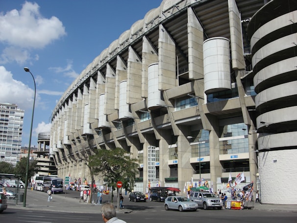 A large, modern sports stadium with a curved roof and prominent architectural features dominates the scene. Surrounding the structure, people and several vehicles are visible, indicating a busy urban environment. Flags and banners suggest an event or gathering. Nearby, urban buildings and trees add to the bustling atmosphere.