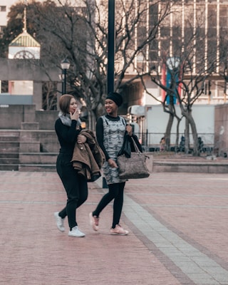 Two people walking together in an urban setting, both smiling and engaged in conversation. One is wearing a black outfit with a grey patterned top, carrying a large bag. The other is in a black jacket and pants, holding a coat. Trees without leaves and a building with colorful columns in the background.