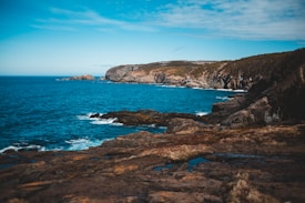 A rugged coastline with rocky cliffs overlooking a deep blue ocean. The sky is partly cloudy, and the water near the shore is a lighter blue due to the shallower depths and white sea foam created by gentle waves. The vegetation on the cliffs appears scarce and rugged, typical of a coastal environment.