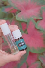 A hand holds a small amber bottle labeled 'Peppermint Essential Oil' alongside a clear glass vial with a white cap. The background features large, vividly colored pink and green leaves.