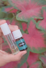 A hand holds a small amber bottle labeled 'Peppermint Essential Oil' alongside a clear glass vial with a white cap. The background features large, vividly colored pink and green leaves.