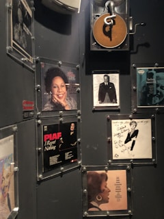 A wall decorated with various framed album covers and autographed photographs. There is a dark wall partially covered with images of musicians and singers. Some frames contain vintage album artworks while others feature black and white portraits with handwritten notes.