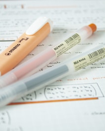 Three pens are placed diagonally on top of handwritten notes on graph paper. One pen is orange and white with the brand name 'STABILO' visible, while the other two pens are transparent with colored inks and 'MUJI' labels indicating they are made in Japan.
