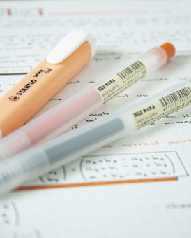 Three pens are placed diagonally on top of handwritten notes on graph paper. One pen is orange and white with the brand name 'STABILO' visible, while the other two pens are transparent with colored inks and 'MUJI' labels indicating they are made in Japan.