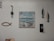 A wall decorated with various nautical-themed items, including a painting of boats on the sea, two hanging fish sculptures, a decorative fishing hook, and a piece of rope shaped like a fish. There is also a small shelf holding a candle.