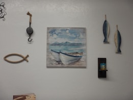 A wall decorated with various nautical-themed items, including a painting of boats on the sea, two hanging fish sculptures, a decorative fishing hook, and a piece of rope shaped like a fish. There is also a small shelf holding a candle.