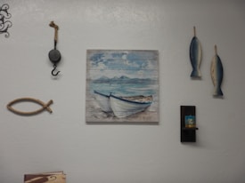 A wall decorated with various nautical-themed items, including a painting of boats on the sea, two hanging fish sculptures, a decorative fishing hook, and a piece of rope shaped like a fish. There is also a small shelf holding a candle.
