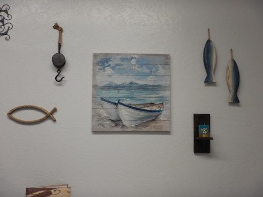 A wall decorated with various nautical-themed items, including a painting of boats on the sea, two hanging fish sculptures, a decorative fishing hook, and a piece of rope shaped like a fish. There is also a small shelf holding a candle.