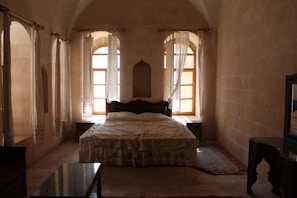 A cozy bedroom features a large bed with an ornate wooden headboard and matching bedside tables, framed by tall, arched windows adorned with sheer curtains that let in soft, natural light. The stone walls and high ceiling add a rustic charm, while a patterned rug lies on the floor at the foot of the bed. A dark wooden desk with a mirror completes the furniture set.
