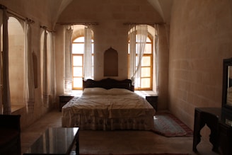 A cozy bedroom features a large bed with an ornate wooden headboard and matching bedside tables, framed by tall, arched windows adorned with sheer curtains that let in soft, natural light. The stone walls and high ceiling add a rustic charm, while a patterned rug lies on the floor at the foot of the bed. A dark wooden desk with a mirror completes the furniture set.