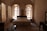 A cozy bedroom features a large bed with an ornate wooden headboard and matching bedside tables, framed by tall, arched windows adorned with sheer curtains that let in soft, natural light. The stone walls and high ceiling add a rustic charm, while a patterned rug lies on the floor at the foot of the bed. A dark wooden desk with a mirror completes the furniture set.