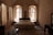 A cozy bedroom features a large bed with an ornate wooden headboard and matching bedside tables, framed by tall, arched windows adorned with sheer curtains that let in soft, natural light. The stone walls and high ceiling add a rustic charm, while a patterned rug lies on the floor at the foot of the bed. A dark wooden desk with a mirror completes the furniture set.