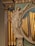 A classical statue of a woman with draped garments, holding a harp in one hand and gesturing upwards with the other. The figure is integrated into an ornate architectural setting featuring columns, decorative floral elements, and gold pipes resembling those of an organ. Blue drapery accents the background.