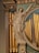A classical statue of a woman with draped garments, holding a harp in one hand and gesturing upwards with the other. The figure is integrated into an ornate architectural setting featuring columns, decorative floral elements, and gold pipes resembling those of an organ. Blue drapery accents the background.