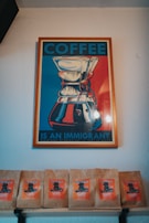 A framed poster with an artistic depiction of a coffee maker, featuring bold colors. The text reads 'COFFEE IS AN IMMIGRANT'. Below the poster, there are several coffee bags displayed on a shelf, each with a vibrant orange label.