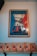 A framed poster with an artistic depiction of a coffee maker, featuring bold colors. The text reads 'COFFEE IS AN IMMIGRANT'. Below the poster, there are several coffee bags displayed on a shelf, each with a vibrant orange label.
