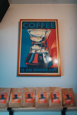A framed poster with an artistic depiction of a coffee maker, featuring bold colors. The text reads 'COFFEE IS AN IMMIGRANT'. Below the poster, there are several coffee bags displayed on a shelf, each with a vibrant orange label.