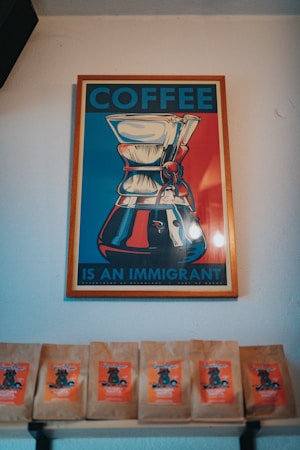 A framed poster with an artistic depiction of a coffee maker, featuring bold colors. The text reads 'COFFEE IS AN IMMIGRANT'. Below the poster, there are several coffee bags displayed on a shelf, each with a vibrant orange label.