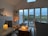 A cozy interior filled with soft lighting from several candles placed on various surfaces around the room. Large windows offer a view of a tranquil outdoor setting with a deck overlooking trees and a lake. The furniture is arranged to create a comfortable sitting area.