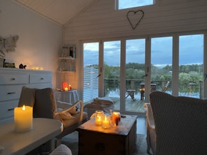 A cozy interior filled with soft lighting from several candles placed on various surfaces around the room. Large windows offer a view of a tranquil outdoor setting with a deck overlooking trees and a lake. The furniture is arranged to create a comfortable sitting area.