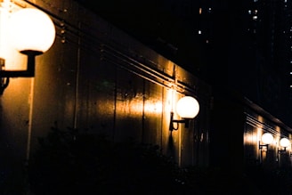 A series of spherical outdoor lights are mounted along a dark wall, casting a warm glow. The lights illuminate the structure, creating subtle reflections and shadows. The background is a dark cityscape with scattered, dimly-lit windows.