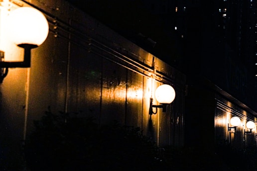 A series of spherical outdoor lights are mounted along a dark wall, casting a warm glow. The lights illuminate the structure, creating subtle reflections and shadows. The background is a dark cityscape with scattered, dimly-lit windows.