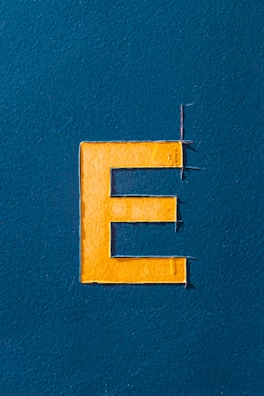 A large uppercase letter 'E' is prominently displayed in a bright yellow color against a textured dark teal background. The letter appears to be cut from a material and slightly raised from the surface, with visible cut marks and slight shadows.