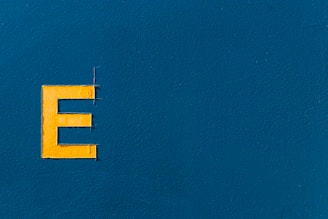 A bold, yellow letter 'E' positioned on the left side of a textured blue background. The surface is smooth with slight variations in the paint texture.
