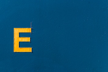 A bold, yellow letter 'E' positioned on the left side of a textured blue background. The surface is smooth with slight variations in the paint texture.