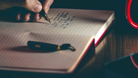 A hand writing with a fountain pen on an open lined notebook. The page shows some text written in cursive. Beside the notebook lies a matching pen cap. The lighting suggests a warm, focused environment, possibly under a lamp.