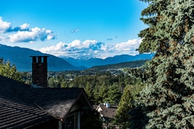 A scenic landscape featuring a residential rooftop with a chimney, set against a backdrop of lush green trees and forested hills. Majestic mountains stretch across the horizon, under a bright blue sky dotted with white clouds.