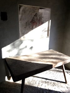 A rustic wooden table with three legs stands in a dimly lit room. Sunlight casts geometric shadows on the textured wall behind it, where a faded artwork or poster is displayed, depicting figures and a building. The atmosphere suggests a vintage or historical setting.