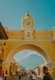 A vibrant street scene features a prominent yellow archway with a clock, typical of Spanish colonial architecture. People are walking beneath the arch, framed by colorful buildings, and a distant volcano with a clear sky overhead.