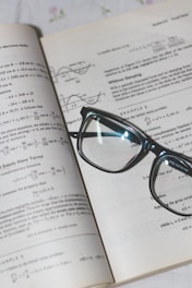 An open book with mathematical formulas and graphs is accompanied by a pair of black-rimmed glasses resting on the pages.