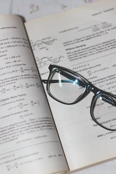 An open book with mathematical formulas and graphs is accompanied by a pair of black-rimmed glasses resting on the pages.
