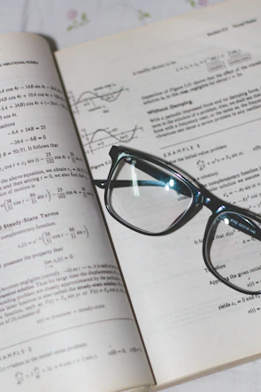 An open book with mathematical formulas and graphs is accompanied by a pair of black-rimmed glasses resting on the pages.