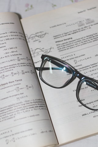 An open book with mathematical formulas and graphs is accompanied by a pair of black-rimmed glasses resting on the pages.