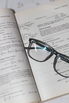 An open book with mathematical formulas and graphs is accompanied by a pair of black-rimmed glasses resting on the pages.