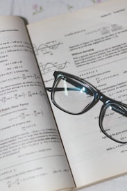 An open book with mathematical formulas and graphs is accompanied by a pair of black-rimmed glasses resting on the pages.