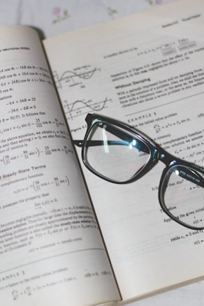 An open book with mathematical formulas and graphs is accompanied by a pair of black-rimmed glasses resting on the pages.