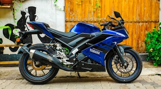 A sleek blue Yamaha motorcycle parked on a paved surface against a wooden backdrop. The bike's design is sporty, with aerodynamic features and a prominent exhaust system. Surrounding the scene are decorative elements such as green plants climbing the wall and a mural-like artwork on the adjacent white wall.