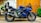 A sleek blue Yamaha motorcycle parked on a paved surface against a wooden backdrop. The bike's design is sporty, with aerodynamic features and a prominent exhaust system. Surrounding the scene are decorative elements such as green plants climbing the wall and a mural-like artwork on the adjacent white wall.