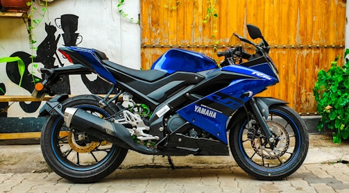 A sleek blue Yamaha motorcycle parked on a paved surface against a wooden backdrop. The bike's design is sporty, with aerodynamic features and a prominent exhaust system. Surrounding the scene are decorative elements such as green plants climbing the wall and a mural-like artwork on the adjacent white wall.