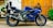 A sleek blue Yamaha motorcycle parked on a paved surface against a wooden backdrop. The bike's design is sporty, with aerodynamic features and a prominent exhaust system. Surrounding the scene are decorative elements such as green plants climbing the wall and a mural-like artwork on the adjacent white wall.