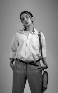 A person stands confidently, wearing a white button-up shirt and tailored trousers. The pose is relaxed, with hands in pockets. The attire is accessorized with a strap bag and a bracelet, and a unique spiral-like earring adds a touch of individuality.
