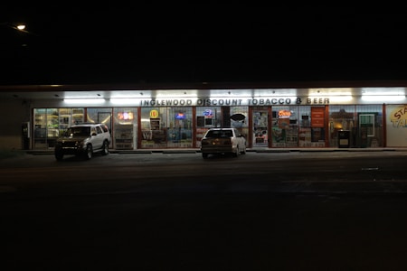 A convenience store is lit up at night with advertisements for cigarettes and beer displayed on the windows. Two parked cars are in the dimly lit parking lot. The store signage reads 'Inglewood Discount Tobacco & Beer.' Bright neon and illuminated signs adorn the windows, giving a vibrant yet subdued atmosphere.