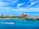 A scenic view of a vibrant waterfront featuring a large resort with multiple pink-colored buildings surrounded by lush greenery and colorful houses. A small white boat is cruising on the clear blue water, while a cruise ship is visible on the distant horizon under a partly cloudy sky.